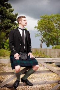 A Scotsman in Highland Dress sitting on a gate outdoors.